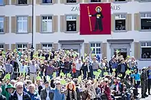 Photographie d'un groupe de femmes et d'hommes, dont certains sur une estrade, tenant en l'air un carton vert ; au fond, un bâtiment sur lequel un drapeau carré représentant un homme tenant un livre et un bâton sur un fond rouge.