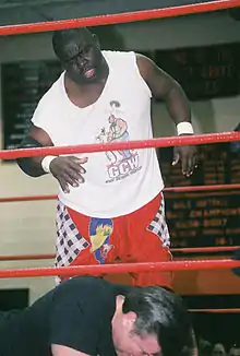 Un catcheur noir avec un t-shirt blanc devant son adversaire au sol.