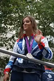Une femme qui porte une médaille d'argent olympique autour du cou, se tient debout derrière une barre métallique et fait une signe de la main.