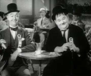 Image extraite d'un film en noir et blanc montrant deux acteurs assis autour d'une tableau, portant chacun un chapeau-melon noir.