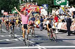 Photographie d'un coureur cycliste portant un maillot rose et levant les bras, devant d'autres coureurs cyclistes.