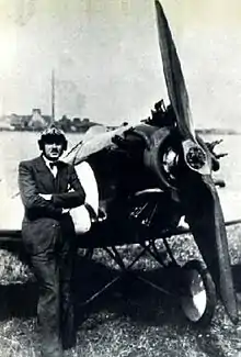 Une photo en noir et blanc d'un homme en costume et nœud papillon, portant un casque de pilote, debout devant un petit monoplan à hélice