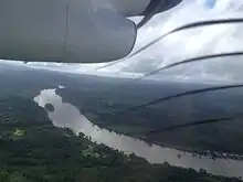 Vue aérienne d'une rivière entièrement bordée d'arbre et de foret dense, avec quelques maisons dans des clairières proches des rives ; au premier plan, un le capot d'un moteur d'avion