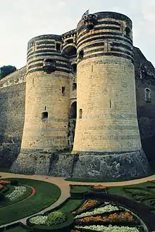 Photographie d'un château fort en schiste et tuffeau. Deux tours imposantes marquent une ancienne porte d'accès.