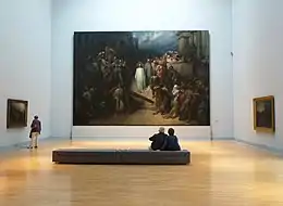 Vue générale d'une salle de musée avec un immense tableau au fond et deux autres sur les murs latéraux. Au premier plan, une banquette où sont assis deux personnages