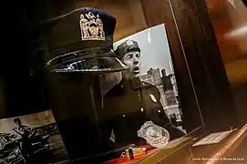 Une casquette, un insigne, un sifflet et deux photos de Louis de Funès dans une vitrine boisée.