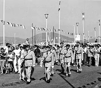 Photo en noir et blanc montrant six gendarmes en uniforme d'apparat défilant sous les regards de touristes, en plein soleil.