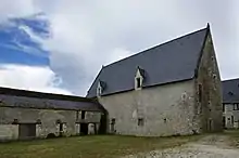 Photographie en couleurs et en trois quarts d'un bâtiment à toiture pentue recouverte d'ardoises munie de fenêtres à meneaux, une seconde construction formant un angle droit visible à gauche.