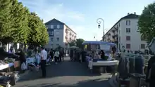 vue d'une rue bordée d'arbres et d'immeubles type HLM de quatre niveaux, au premier plan étals d'une brocante et badauds