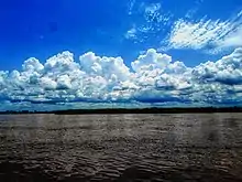Une large coulée d'eau noire ridée de quelques vaguelettes passe sous un ciel bleu et des nuages blancs.