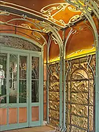 Photo en couleur d'un coin de pièce aux tons chauds dont les murs en relief tarabiscotés sont encadrés d'arabesques en fer forgé
