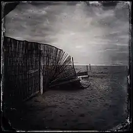 photographie en noir et blanc représentant une plage déserte avec à gauche une clôture légère s'afaissant sur elle-même.