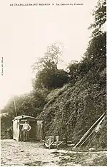 Carte postale en noir et blanc : photographie d'un monsieur debout, devant une cabane marquée « passeur », ayant en main une grande barre de bois de plusieurs mètres de longueur
