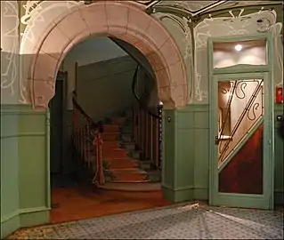 Photo en couleur d'un espace avec une ouverture en arc de cercle donnant sur un escalier et une porte vitrée surmontée d'arabesques peintes