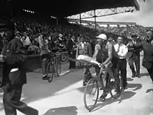 Photographie en noir et blanc d'un cycliste accomplissant un tour d'honneur, un bouquet à la main, devant une foule enthousiaste.