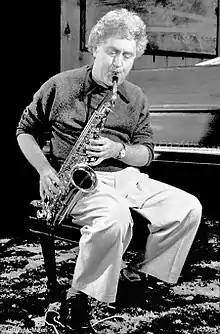 Lee Konitz, vêtu d'un pantalon clair et d'un pull sombre, est assis sur le tabouret d'un piano. Tournant le dos au clavier, il joue de son saxophone alto. La photo est en noir et blanc.