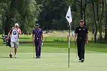 De gauche à droite, sur un green de golf : un golfeur habillé de noir, un club à la main ; un piquet muni d'un drapeau, 2 balles sont à proximité ; un golfeur habillé de violet, un club à la main ; Un homme en short, habillé de blanc, portant un sac de clubs de golf