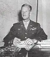 Photographie en noir et blanc d'un officier assis sur un bureau et vêtu d'un costume militaire sombre