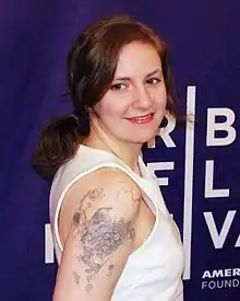 Portrait en buste d'une femme aux cheveux châtains attachés vers l'arrière, regardant par-dessus son épaule droite. Elle porte un haut blanc sans manche, rendant visible un tatouage sur son bras droit, représentant un animal sortant la tête d'un champ de fleurs.