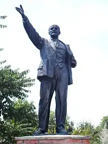 Photo couleur, vue par dessous, d'une statue d'un homme en costume, debout, de face, le bras droit levé devant lui, paume de la main ouverte (ciel nuageux blanc en arrière-plan).