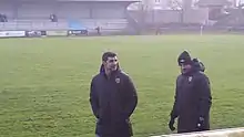 Photo de deux hommes debout devant un terrain de football. Au fond se trouve une tribune de stade.