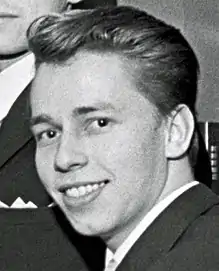 Photographie en noir et blanc d'un homme de peau blanche, brun, de trois-quarts, souriant, regardant l'objectif, en gros plan.