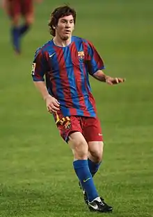 Photographie couleur. Lionel Messi foulant une pelouse. Il revêt une tenue de match aux couleurs du FC Barcelone.