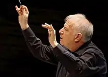 Photographie en couleurs d'un homme aux cheveux blancs vu de profil gauche, vêtu d'une chemise noire et tenant une baguette de chef d'orchestre de la main droite, se détachant sur un fond noir uniforme