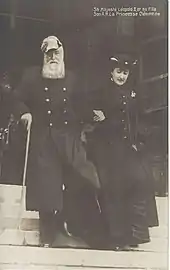 Photo de Léopold II en tenue de lieutenant-général, portant une barbe blanche et coiffé d'un bicorne, s'appuyant de la main droite sur une canne et donnant le bras gauche à sa fille Clémentine, vêtue d'un long manteau sombre et coiffée d'un petit chapeau.Tous deux descendent un escalier.