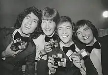 Quatre jeunes hommes tout sourire en smoking, tenant chacun un appareil photo.