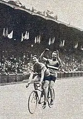 Photo de deux hommes pédalant sur un tandem devant les tribunes d'un stade.