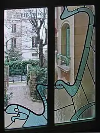 Photo en couleur d'une fenêtre en verre blanc traversé d'arabesques en vitrail vert, donnant sur une petite cour avec un peu de verdure
