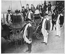 Une large cloche est accrochée sur un chariot entouré par des soldats en uniformes de la guerre d'indépendance.