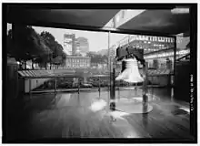 Photographie de la cloche dans un bâtiment moderne en bois et en métal devant une grande baie vitrée où l'on peut voir l'Independence Hall.