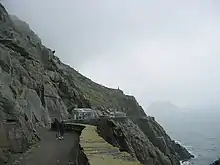 Photographie en couleurs d'un chemin taillé à flanc de falaise et délimité à droite par un parapet en pierre, un bâtiment installé à son extrémité.