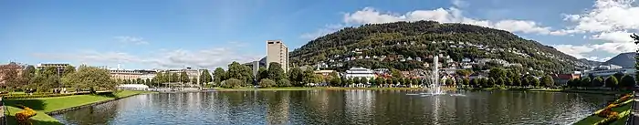 Un lac urbain avec un jet d'eau au milieu, une colline ronde derrière, quelques arbres et immeubles autour.