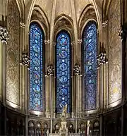 Photographie en couleurs d’une chapelle avec trois vitraux avec de part et d'autre une mosaïque murale et en bas la statue de Jeanne d'Arc.