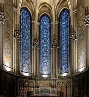 Photographie en couleurs de la chapelle vue de face avec au centre trois vitraux, de part et d'autre une mosaïque murale et en bas le retable.