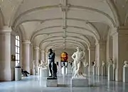 Vue de la galerie dans sa longueur, rythmée de lourd piliers de pierre sous une voute cintrée, au premier plan deux statues d'hommes nus.