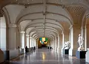 Vue du hall dans sa longueur, rythmé de lourd piliers de pierre sous une voute cintrée parsemée de lumignons, à droite des statues, à gauche des guichets, au fond un lustre multicolore.