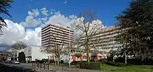 Photographie en couleur représentant, derrière un voile d'arbre sans feuilles, à droite un long bâtiment de huit étages percé de fenêtres carrés, au premier plan un immeuble bas à deux niveaux, plus loin derrière, un immeuble d'une douzaine d'étages rayé de rouge