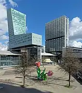 Photographie en couleur d'une vaste place minérale avec en son centre une sculpture représentant des tulipes colorées encadrée de deux arbres sans feuilles et, derrières, deux tours, la première, à gauche, en forme de chaussure de ski, la seconde, plus loin à droite, de forme parallélépipédique