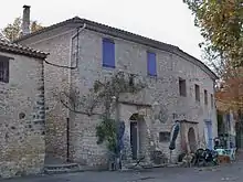 Maison à un étage en moellons, murs clairs, volets violets fermés. Une glycine pousse contre la façade.