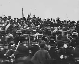 Photographie en noir et blanc montrant une foule d'hommes : face à lui, de dos, se tiennent de nombreux soldats et spectateurs.