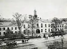 A black and white photograph, showing a stately-looking two-storey building with white walls, extending out of shot to the left and right, with an arched cart entrance at the centre. A modest clocktower rises above the entrance, and the building is surrounded by neat shrubbery and iron railings. A wide street crosses left-right outside of the fence, with a handful of horse-drawn carts and pedestrians in 19th-century clothing.