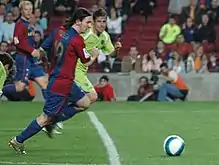 Photographie couleur. Au premier plan, Lionel Messi court pour le FC Barcelone sur un terrain. Il dispute un ballon à la lisière de la surface de réparation avec un adversaire revêtant les couleurs du Getafe FC, situé au second plan. À l’arrière plan, une tribune accueillant un public clairsemé.