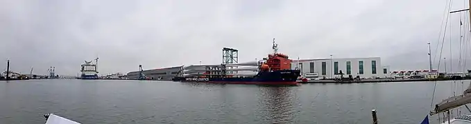 Livraison des pâles d'éoliennes en mer dans le port de Saint Nazaire. Au second plan, le navire Vestvind est à quai, chargé des pales d'éoliennes qui seront montées sur les turbines des éoliennes offshore. À l'arrière plan à gauche, le paquebot Celebrity Beyond en cours d'armement dans le bassin de Penhouët. À l'arrière plan à droite, l'usine de moteurs thermiques Man, et à l'extrême droite les turbines entreposées en attendant de repartir vers le champ en mer.