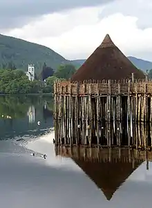 A small, brown conical structure sits on top of wooden piers set into a body of water. Ducks paddle through the water and in the near background there is a tree-lined shore with a white square tower showing amongst the trees. Tree covered hills and grey skies dominate the far background.
