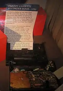 Photo d'une radio-cassette ouverte contenant une fausse bombe entourée de fils et de circuit imprimé, avec un papier d'explication juste derrière.