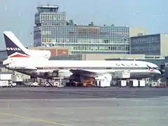 Photo couleur de l'avion, caractérisé par ses trois moteurs, dont deux sont situés sous ses ailes et le troisième sur sa queue. L'avion est peint en blanc et une fine ligne bleue sur le milieu et l'inscription Delta sur l'avant du fuselage. Il se trouve à une porte d'embarquement et des bâtiments de l'aéroport sont visibles à l'arrière-plan.